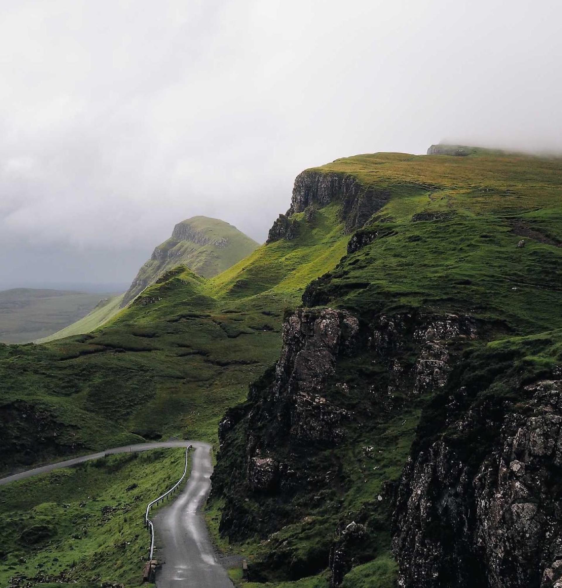 A winding road curves through the Quiraing on the Isle of Skye, Scotland, surrounded by dramatic green, mossy hills under a cloudy, misty sky.