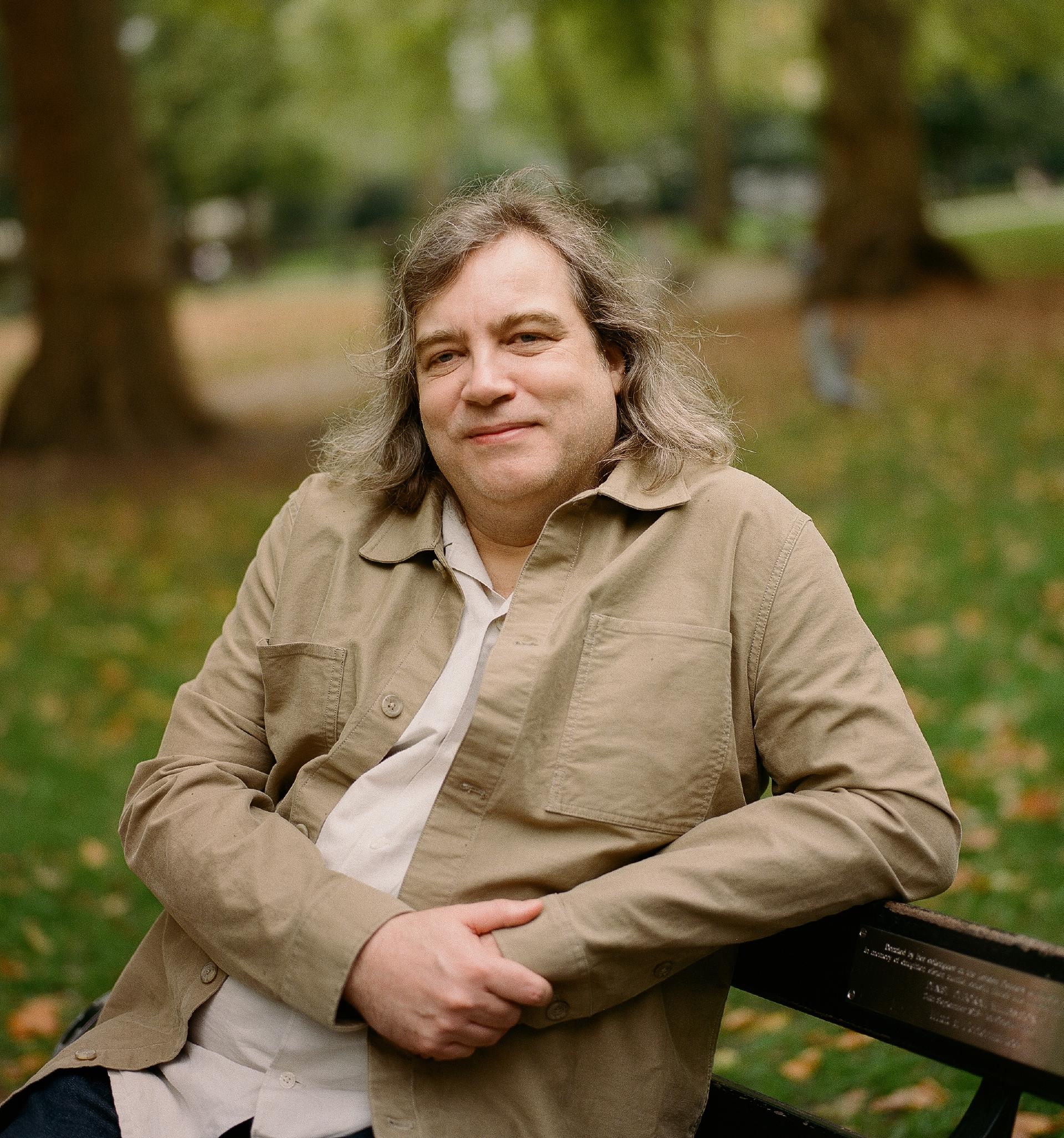 A man with long, ash-toned hair and a khaki jacket sits on a park bench. He looks directly at the camera with a mild, relaxed expression. The background shows trees and the ground is covered with fallen autumn leaves.