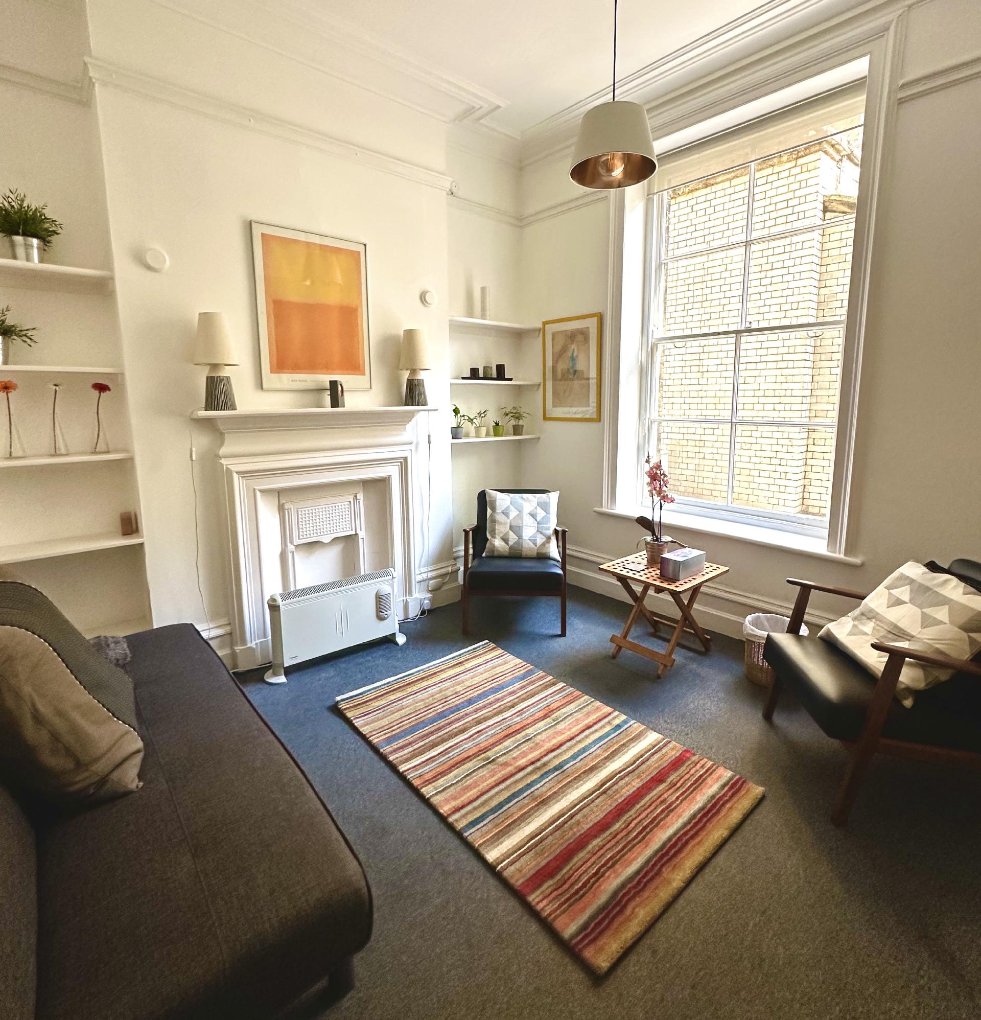 A brightly lit, contemporary room with a gray sofa on the left, a white decorative fireplace flanked by shelves, and a colorful striped rug on the floor. Two dark armchairs and a large window facing a brick wall are also visible.