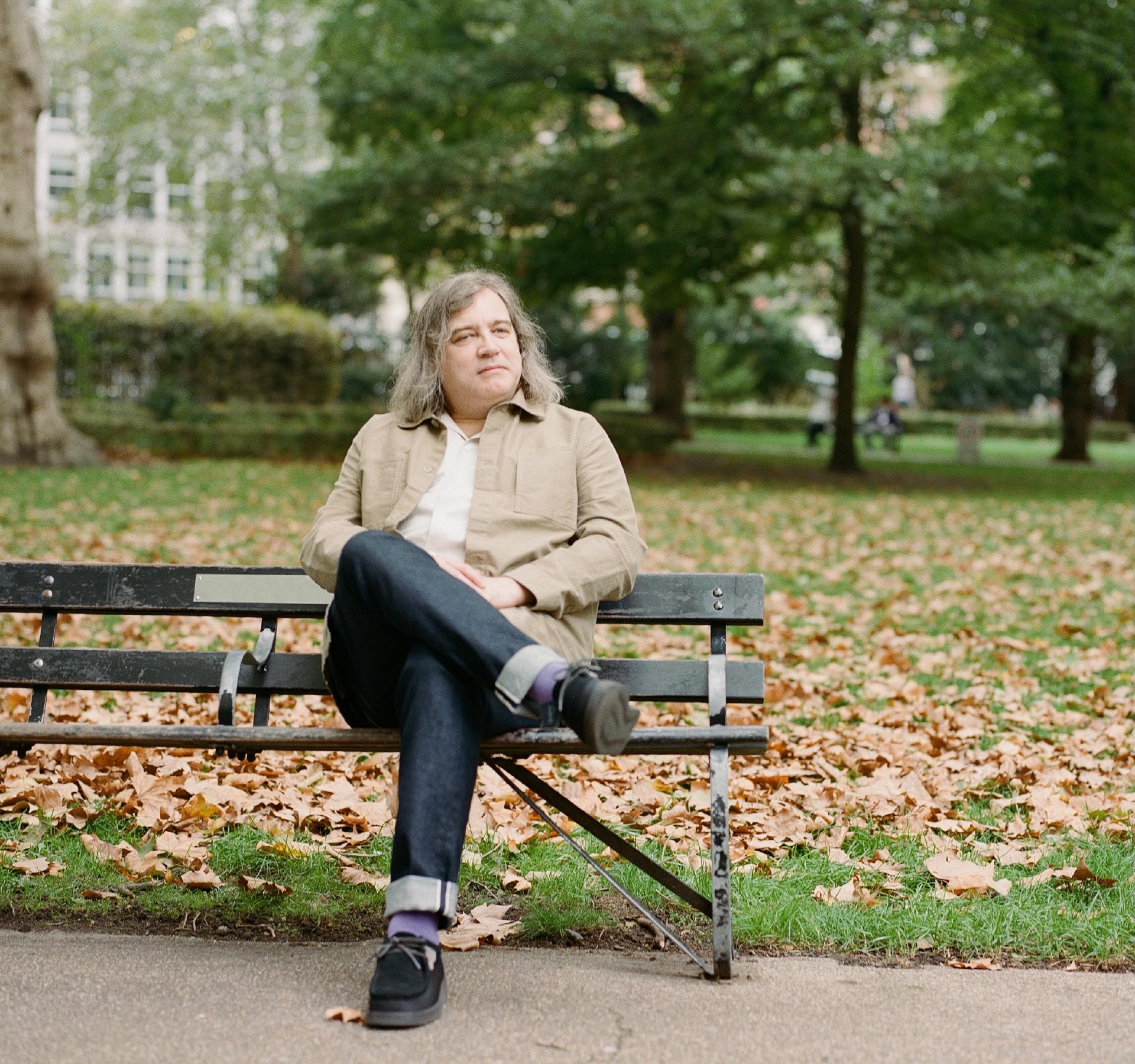 A person with long, wavy hair sits calmly and contemplatively on a park bench in autumn, wearing a tan jacket and dark jeans, with fallen brown leaves scattered on the grass around them.